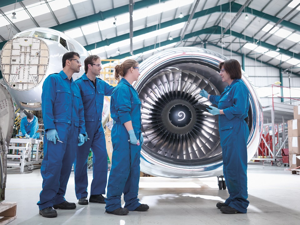 Four mechanics stand in front of a large aircraft turbine.